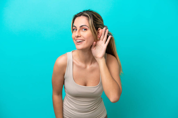 Junge kaukasische Frau isoliert auf blauem Hintergrund hört etwas, indem sie die Hand auf das Ohr legt - Foto, Bild