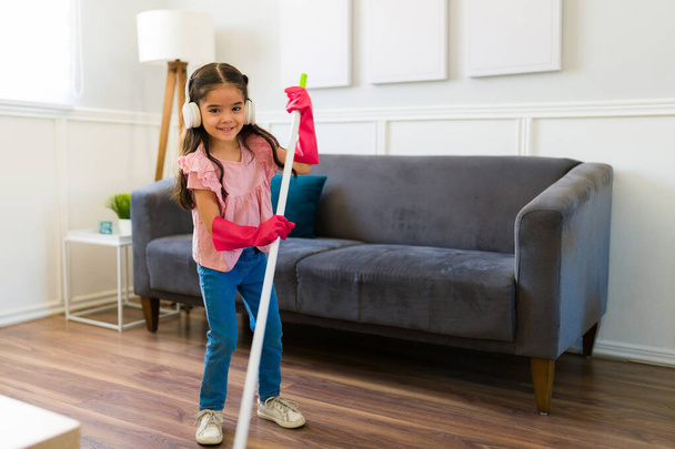 Beautiful young kid smiling while sweeping the living room and helping her family to do housework  - Foto, imagen
