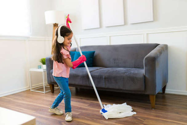 Cheerful young girl with headphones using a broom and cleaning the floor while doing housework - Foto, imagen