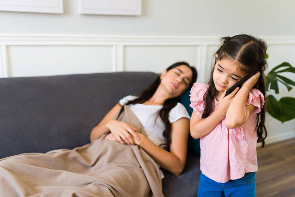 Worried little girl calling a doctor on the phone and asking for help because her sick mother is in pain lying on the sofa at home  - Photo, Image