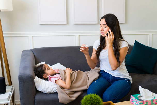 Upset young mom feeling very worried while calling a doctor on the phone because her little daughter is very sick at home - 写真・画像