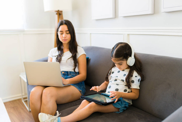 Elfoglalt fiatal anya dolgozik otthonról, és gépel a laptopon a fiatal gyerek játszik videojátékok a táblagépen  - Fotó, kép