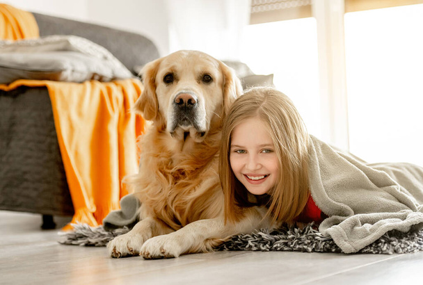 Cheerful child girl lies with golden retriever dog under plaid. Happy preteen girl with purebred doggy labrador spend time on floor. - Фото, зображення