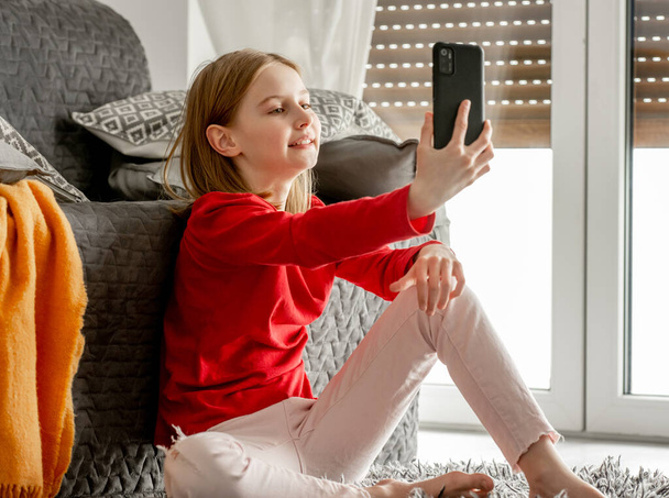 Happy child girl making selfie with smartphone at home. Cheerful child with cellphone sitting on floor and taking photos of herself with mobile camera - Valokuva, kuva