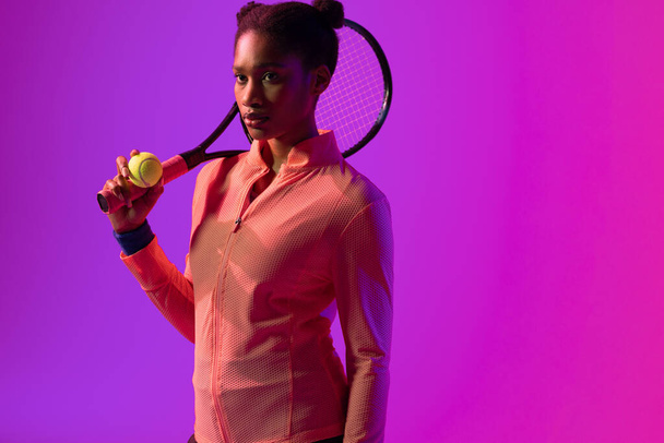 Image of african american female tennis player in violet and pink neon lighting. Sport, movement, fitness and active lifestyle concept. - Photo, image