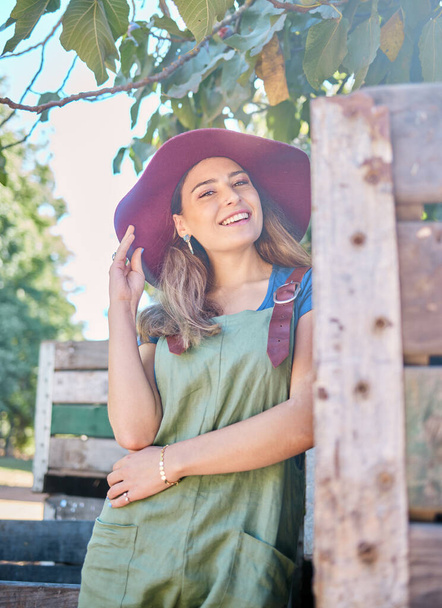Portrait of a woman in a straw hat standing under a tree next to a rustic wooden crate. One young happy female wearing a summer hat and dungaree dress in a garden on a sunny day picking apples. - Foto, Imagen
