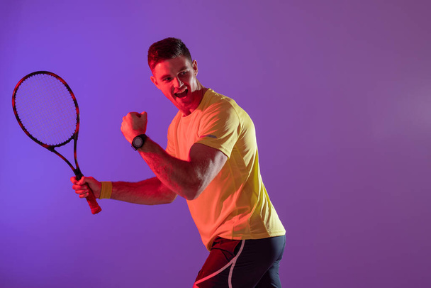 Happy caucasian male tennis player with tennis racket over neon pink lighting. Sport, movement, training and active lifestyle concept. - Foto, afbeelding