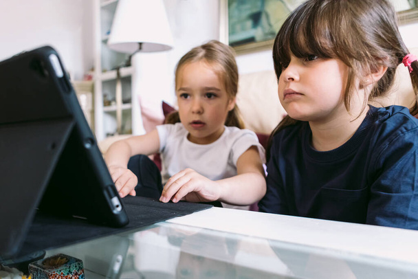 Twee kleine meisjes kijken aandachtig naar een video op een tablet in hun woonkamer. Begrip jeugd, technologie, leren, plezier hebben, internet en connectiviteit. - Foto, afbeelding