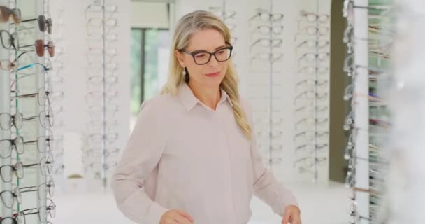 Confident mature woman and owner choosing or selling glasses at an optics store. Portrait of a female optician or optometrist smiling and standing with her arms crossed between shelves in her shop. - Πλάνα, βίντεο