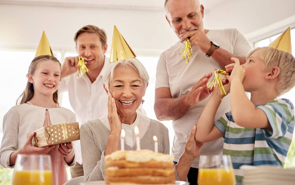 Senior woman celebrating her birthday with family at home, wearing party hats and blowing whistles. Grandma looking at birthday cake and looking joyful while surrounded by her grandkids and and son. - Photo, image