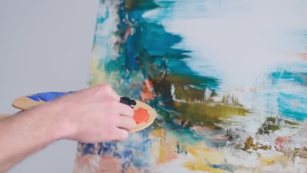 Foto ravvicinata della mano di un artista maschio che tiene un pennello e dipinge un dipinto ad olio. Pittura ad olio colorata ed emotiva. L'artista utilizza vernici blu e arancioni. L'artista dipinge un paesaggio. - Filmati, video