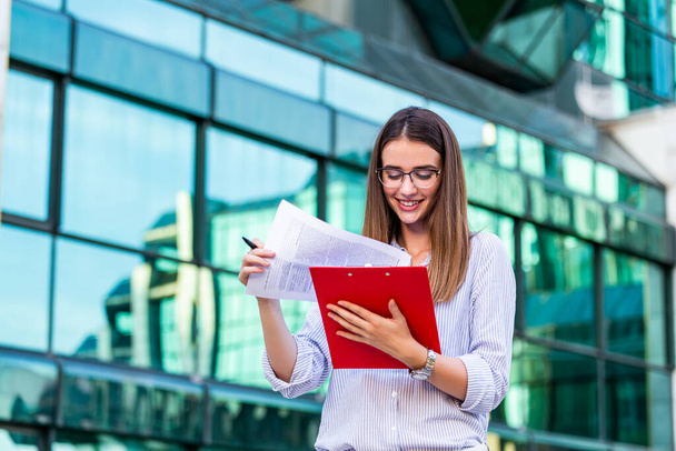 Happy beautiful business woman with clipboard. Portrait Of Successful Business Woman Holding clipboard. Photo business woman wearing suit and holding documents in hands. Panoramic windows background. - Foto, Bild