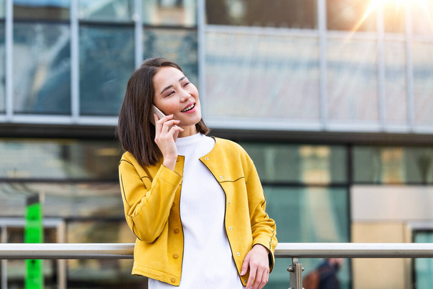Счастливая улыбающаяся деловая женщина разговаривает по мобильному смартфону на улице с офисными зданиями на заднем плане. Представительница бизнеса азиатка разговаривает на мобильном смартфоне - Фото, изображение