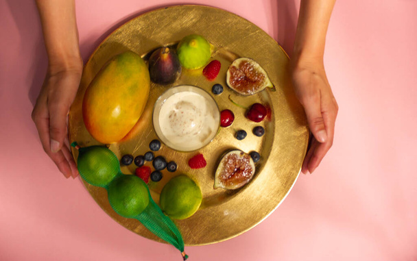 Kadın elleri güzel servis edilmiş bir tabak sunar. Pembe masa manzaralı taze tropikal meyve tabağı. Masada servis yapan yüzü olmayan kız. İncir, mango, çilek, kiraz, limon, tepside dondurma - Fotoğraf, Görsel