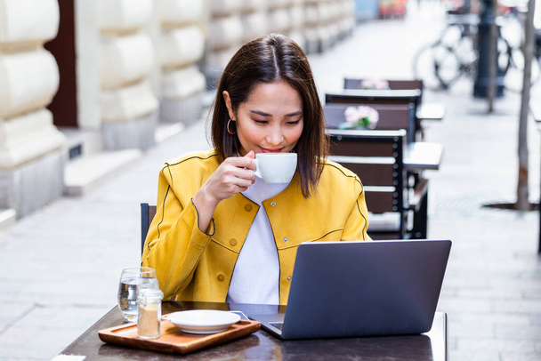 Jeune femme d'affaires asiatique assise à table et prenant des notes dans un carnet. Sur la table se trouve un ordinateur portable, un smartphone et une tasse de café. Apprentissage étudiant en ligne. Blogueur. - Photo, image