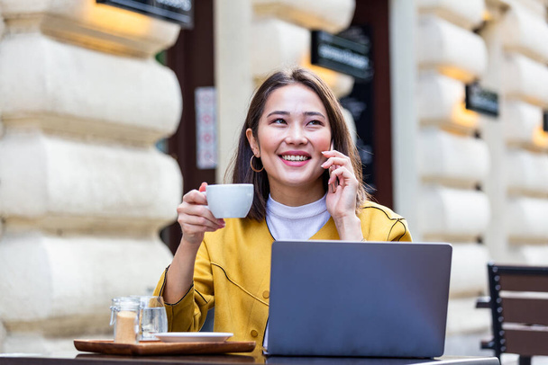 Jeune femme asiatique assis dans un café à la table en bois, boire du café et en utilisant smartphone.On table est ordinateur portable. Fille naviguant sur Internet, bavarder, blogging. - Photo, image