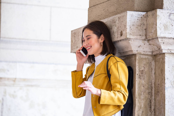 Asian cheerful young woman walking along the street and calling on phone. Asian woman walking on the street, wearing cute trendy outfit and talking om her smart phone. - 写真・画像