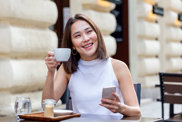 Hymyilevä aasialainen nainen juo kahvia ja käyttää matkapuhelintaan. Tyytyväinen nainen nauttii kupillisesta kahvia. Lähikuva muotokuva kaunis tyttö juo kahvia valkoinen muki kahvilassa - Valokuva, kuva