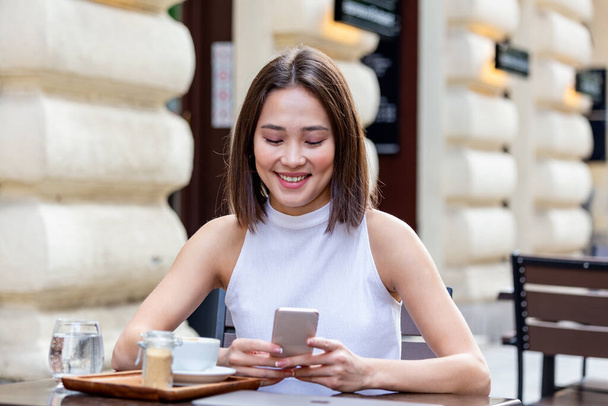 Hymyilevä nainen juo kahvia ja käyttää matkapuhelintaan. Tyytyväinen nainen nauttii kupillisesta kahvia. Lähikuva muotokuva kaunis tyttö juo kahvia valkoinen muki kahvilassa - Valokuva, kuva