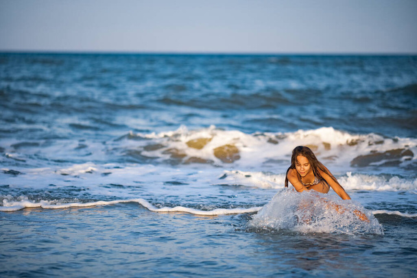 Fat dark-haired pensive girl with dark loose wet hair walks along the blue shallow sea with small waves and enjoys the warm summer sun - Φωτογραφία, εικόνα