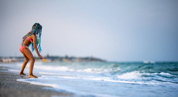 Funny slender cheerful girl playing with small waves, kicking their feet and spinning in place under the warm bright summer sun enjoying the long-awaited vacation - Foto, afbeelding