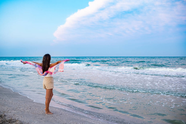 A fat dreamy dark-haired dark-skinned girl in snow-white summer T-shirt and beige shorts is walking along sea sandy beach, looking at horizon and holding bright shawl in hands - Фото, зображення