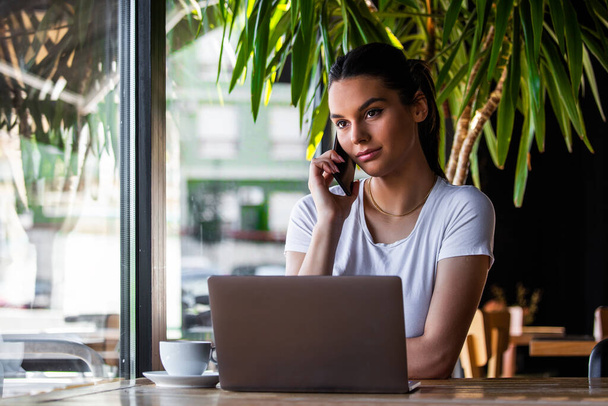 Mladá žena s šálkem kávy a notebookem mluví na svém chytrém telefonu. Žena si dává pauzu. Podnikání Z kavárny - Fotografie, Obrázek