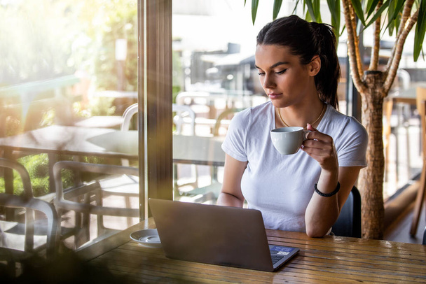 Young beautiful girl sitting at a table in a cafe by the window, drinking cappuccino coffee. Beautiful young working with laptop from coffee shop. Attractive woman sitting in a cafe with a laptop - Fotó, kép
