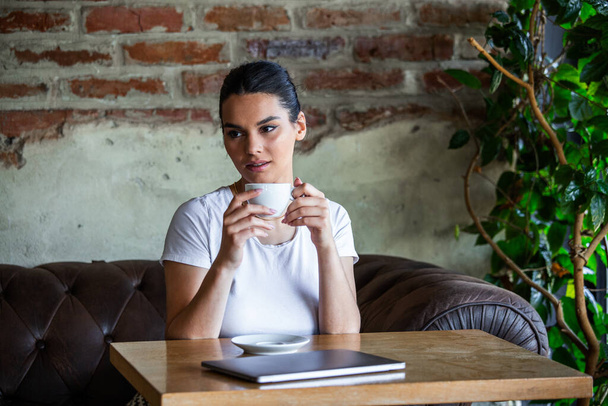 Ευτυχισμένη νεαρή γυναίκα χαλαρώνει στον άνετο καναπέ και χρησιμοποιεί φορητό υπολογιστή σε καφετέρια. Νεαρή γυναίκα απολαμβάνει ένα φλιτζάνι καφέ. Γυναίκα πίνει καφέ και χρησιμοποιεί φορητό υπολογιστή - Φωτογραφία, εικόνα