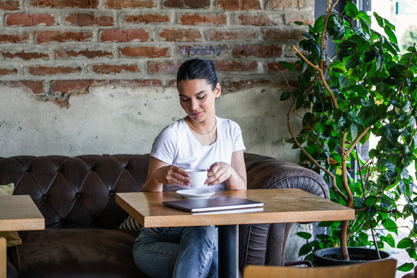 Happy young woman is relaxing on comfortable couch and using laptop at coffee shop. Young woman enjoying a cup of coffee. Woman drinking coffee and using laptop - Foto, afbeelding
