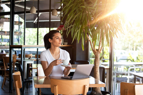 Ένα νεαρό όμορφο κορίτσι κάθεται σε ένα τραπέζι σε ένα καφέ δίπλα στο παράθυρο, πίνοντας καπουτσίνο καφέ. Όμορφη νεαρή που εργάζονται με φορητό υπολογιστή από την καφετέρια. Ελκυστική γυναίκα κάθεται σε ένα καφέ με ένα φορητό υπολογιστή - Φωτογραφία, εικόνα