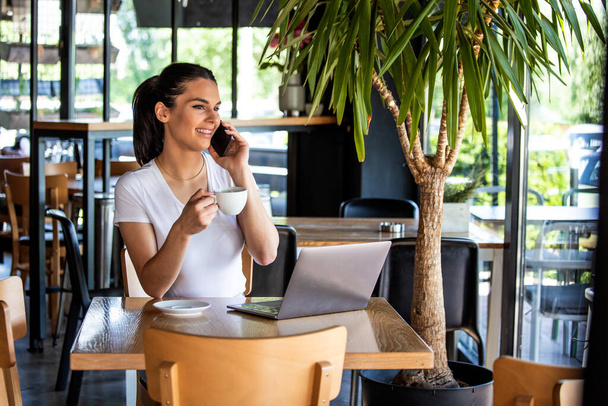 Молода жінка з чашкою кави і ноутбуком говорить на своєму смартфоні. Жінка робить перерву. Ведення бізнесу Від кав'ярні
 - Фото, зображення