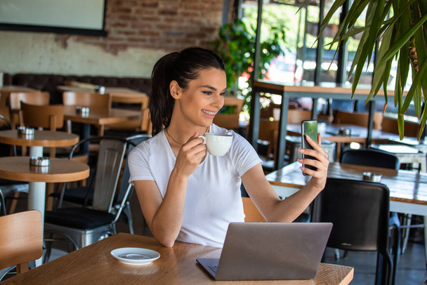 Porträt eines verspielten jungen Mädchens, das ein Selfie mit dem Handy macht, während es mit dem Laptop in einem Café im Freien sitzt - Foto, Bild