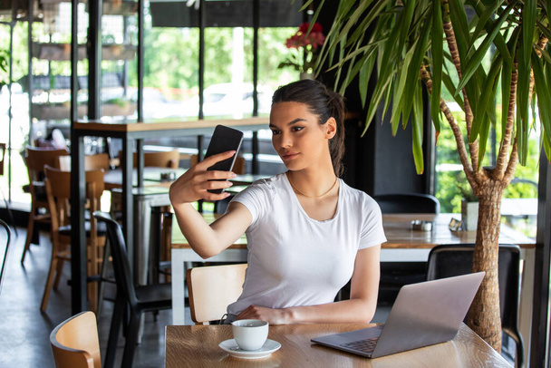 Retrato de una joven juguetona tomando selfie con teléfono móvil mientras está sentada con una computadora portátil en un café al aire libre
 - Foto, imagen