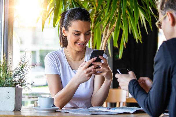 One-on-one meeting.Two young business women sitting at table in cafe. Girl shows colleague information on laptop screen. Girl using smartphone, blogging. Teamwork, business meeting. Freelancers working. - Foto, Bild