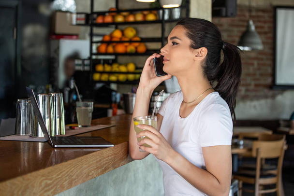 Young charming woman calling with cell telephone while sitting alone in coffee shop during free time, attractive female with cute smile having talking conversation with mobile phone while rest in bar - Photo, image