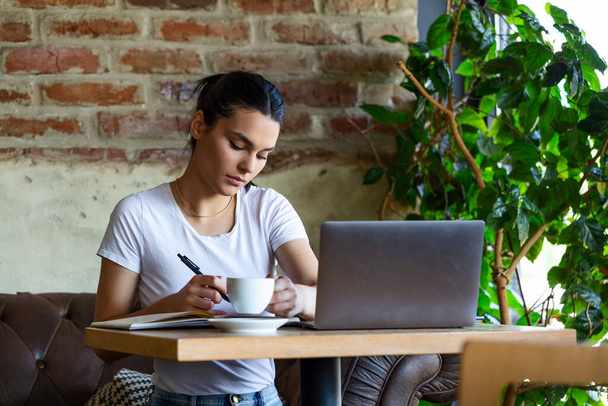 Piękna młoda dziewczyna pracująca w kawiarni z laptopem i robiąca notatki. Kobieta-freelancer łącząca się z internetem przez komputer. Biznes kobieta kryty z kawy i laptopa robi notatki - Zdjęcie, obraz