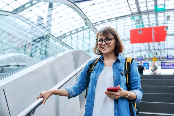 Middle-aged woman with backpack smartphone in her hands walking up stairs, near escalator in modern station building. Urban architecture, urban lifestyle, people concept - Photo, image