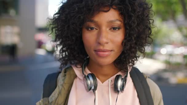 Porträt einer trendigen jungen schwarzen Frau mit unbeschwerter und cooler Attitüde, die in der Stadt steht. Schöne, selbstbewusste und selbstbewusste afrikanische Studentin mit lockigem Afro-Haar und Kopfhörer in der Stadt. - Filmmaterial, Video