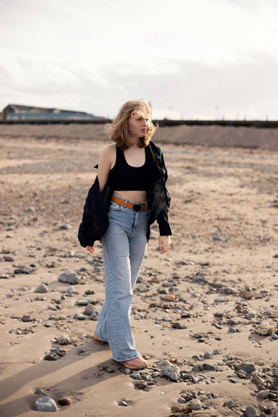 A young blonde girl dressed in a black silk shirt and blue jeans is walking along the beach and enjoying the cloudy weather - Foto, Bild