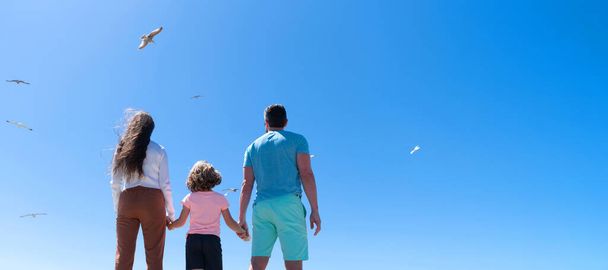 Family of mom father and child looking in future hold hands in summer, back view, summer vacation. Banner of family embracing on sea beach. Summer family vacation, horizontal banner - 写真・画像