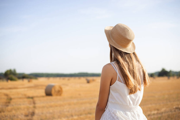 Lonely girl in a white dress, straw hat sitting on a bale in the agricultural field after harvesting. Beautiful girl in the field - Foto, immagini