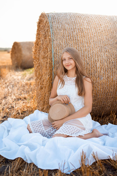 Portrait of a young girl sitting on a white cloth near the bale of straw in agricultural field. Beautiful girl on a farm field after the harvest - Фото, изображение
