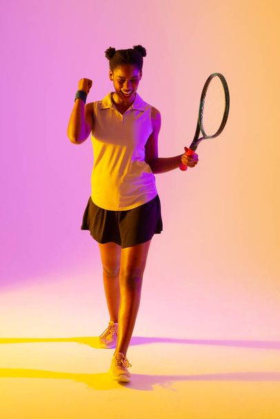 Vertical image of successful african american female tennis player in violet and yellow lighting. Sport, movement, fitness and active lifestyle concept. - Zdjęcie, obraz
