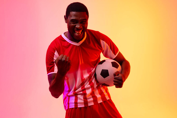 Portrait of happy african american male soccer player with football over neon pink lighting. Sport, movement, training and active lifestyle concept. - Foto, imagen