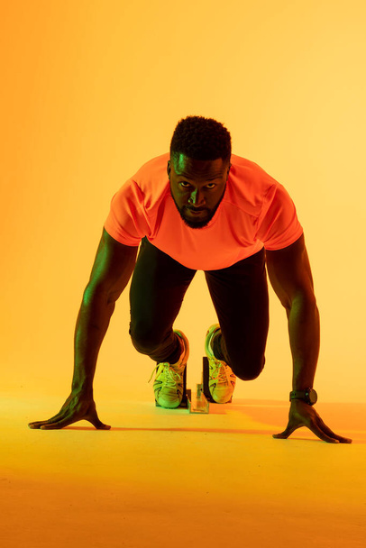 Vertical image of african american male athlete preparing for run in yellow lighting. Sport, movement, fitness and active lifestyle concept. - Foto, Bild