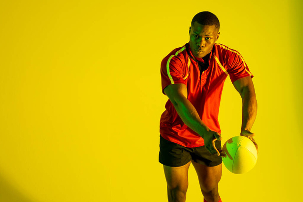 African american male rugby player with rugby ball over yellow lighting. Sport, movement, training and active lifestyle concept. - Foto, Imagen