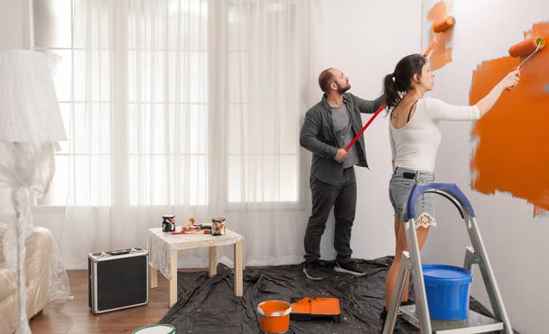 Confident couple painting room with orange color paint to redecorate apartment with paintbrush and roller. Doing diy manual renovation using decorating tools and equipment, housework change. - Foto, afbeelding