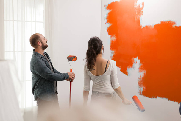 Man and woman painting with orange color, using paint to redecorate walls in apartment. Renovating house interior with paintbrush, renovation tools and roller, decorating room. - Foto, afbeelding