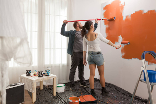 Pareja feliz pintando pared con pintura naranja, redecorando apartamento con nuevo color. Renovación y mejora de casas con pincel, rodillo manual, renovación de paredes de la sala de estar. - Foto, imagen